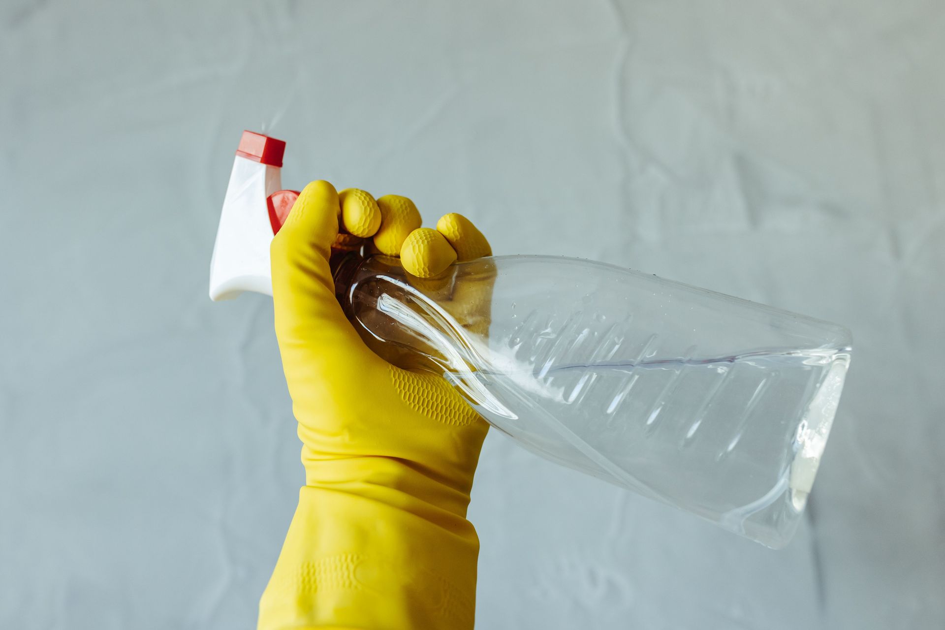 Close-up of a hand wearing yellow gloves holding a transparent spray bottle, isolated on a plain background.